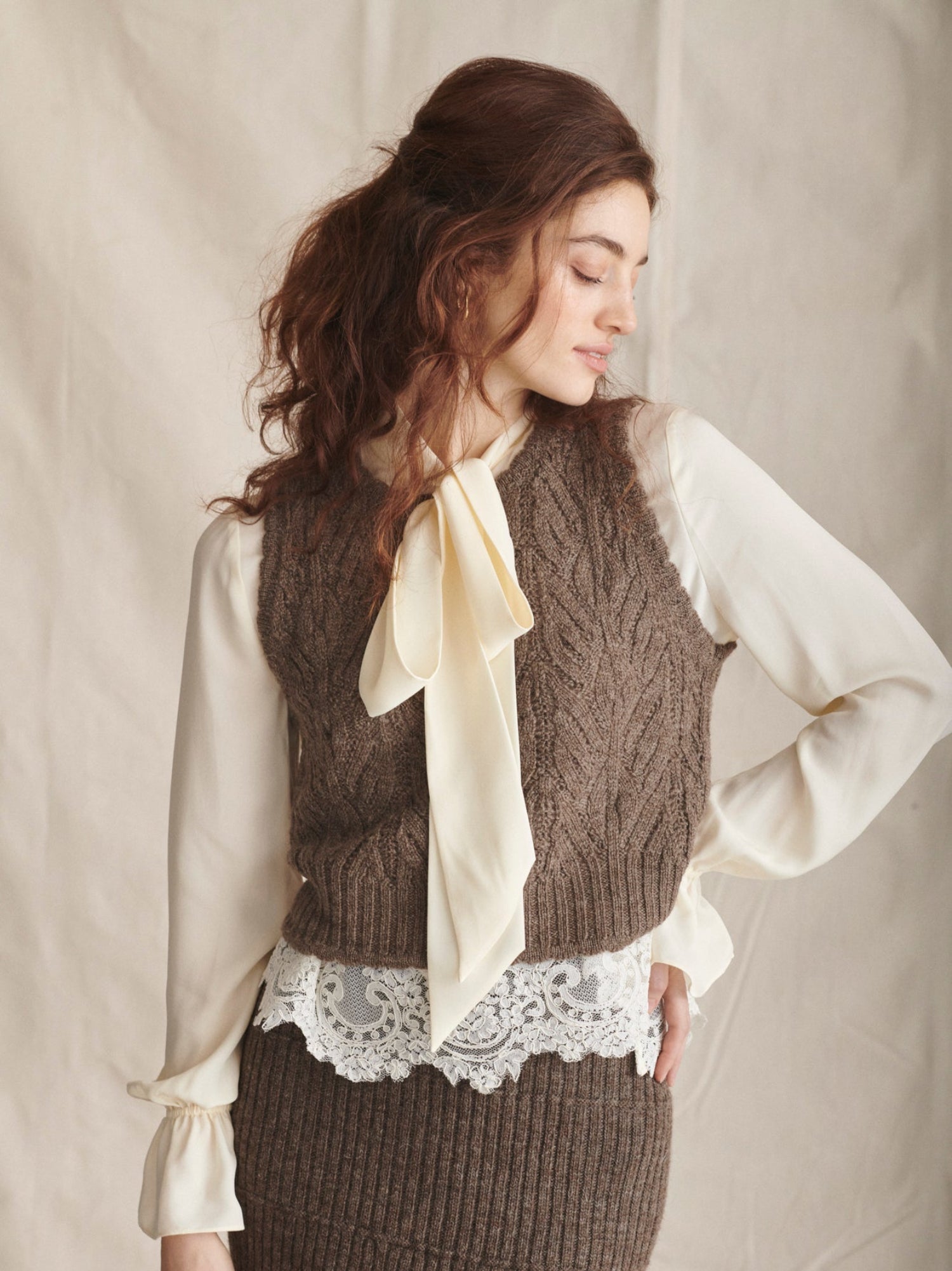 Woman wearing a brown knitted vest over a white blouse with a beige scarf, against a plain background.